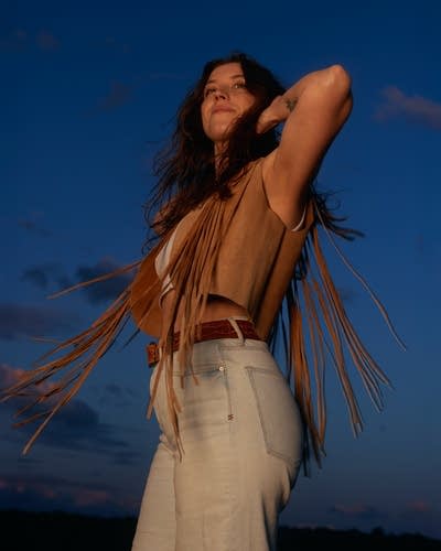 Clare Doyle wears a brown vest, white tank, light-colored denim. A dramatic blue sky is behind her.