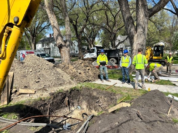 A group of construction workers in hi-vis vests stand next to an open hole in a yard.