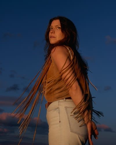 Clare Doyle wears a brown vest, white tank, light-colored denim. A dramatic blue sky is behind her.