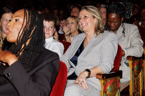 A woman in a gray suit smiles while sitting in a theater seat.