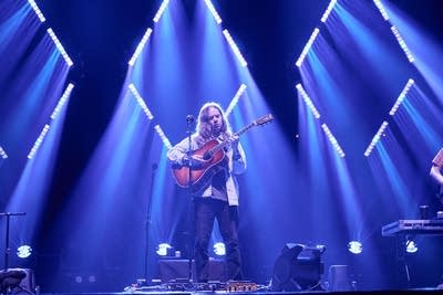 A musician plays flatpick guitar onstage in a large arena