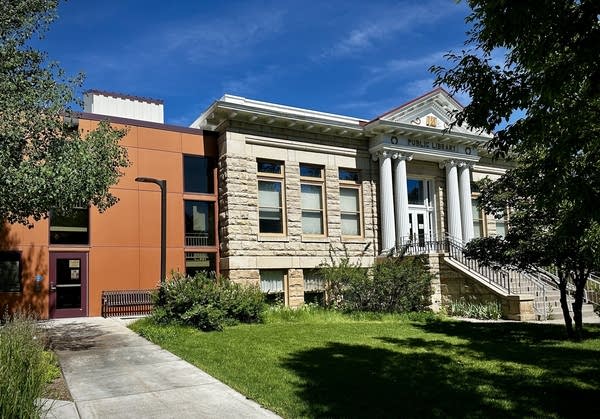 An older building with white pillars attached to a newer, brown-colored building. Green grass and trees surround it.