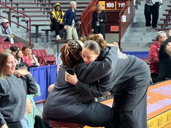 Two women hug on the sideline of a basketball court