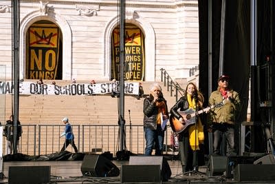Three musicians perform together on an outdoor stage at a large rally