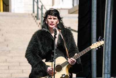 A musician sings and plays guitar on an outdoor stage at a large rally