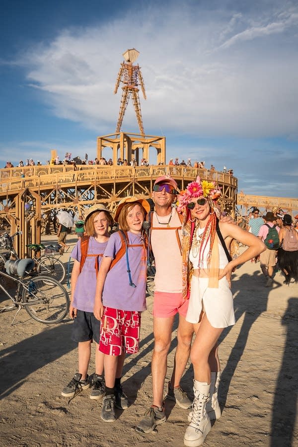 A family of four poses for a photo at the Burning Man festival. A large wooden sculpture of a person is seen in the distance.