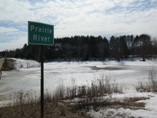 A sign reading "Prairie River" stands next to the ice-covered waterway.