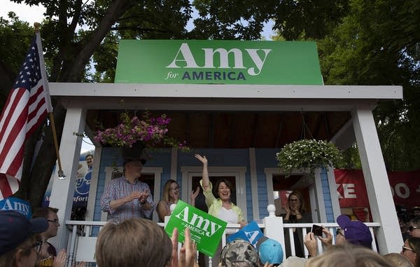 A woman waves to a crowd as she stands on a white porch.