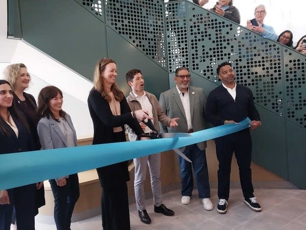 A woman stands next to the mayor with giant scissors cutting a light blue ribbon.