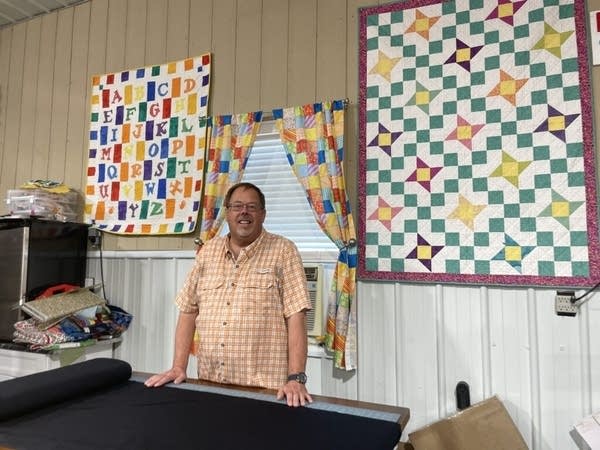 An older man, wearing glasses and a checkered shirt, stands at a table. Black fabric is laid out on the table, ready to be measured and cut. Colorful quilts hang on the wall behind him.