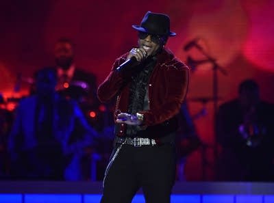 A man in sunglasses, a jacket and fedora sings onstage