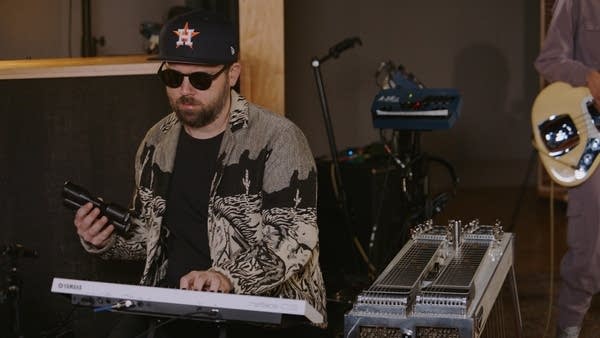 A musician simultaneously plays a synthesizer and a shaker in a recording studio