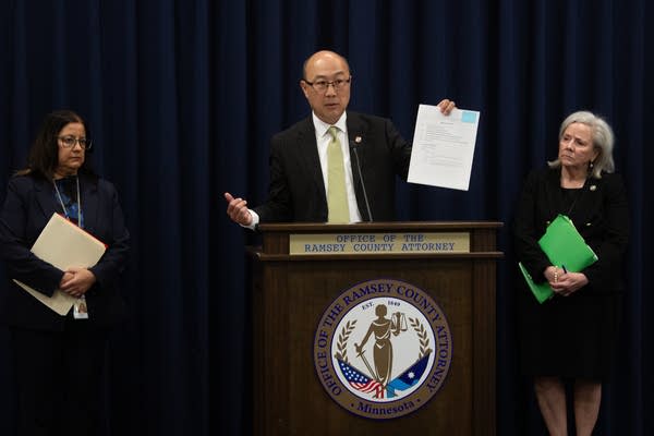 A man at a podium holds up a document while flanked by two women.