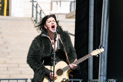 A musician sings and plays guitar on an outdoor stage at a large rally
