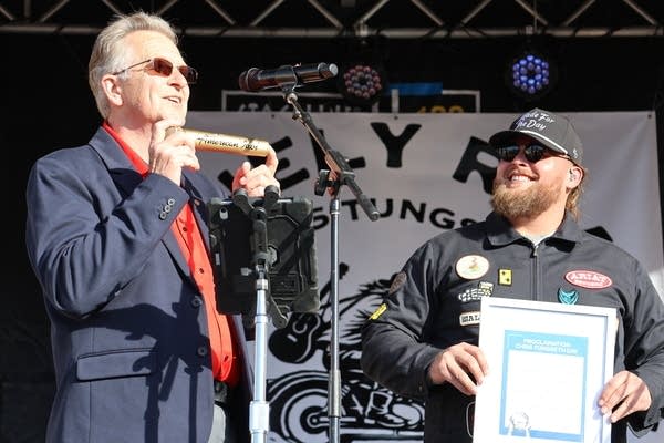 A man in a suit presents a golden microphone to another man holding a framed plaque.