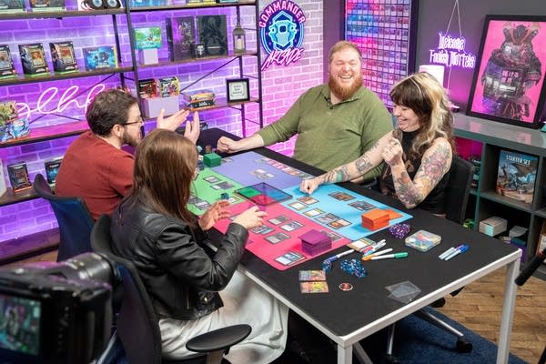 Four people sit on a stage to play a game of cards.
