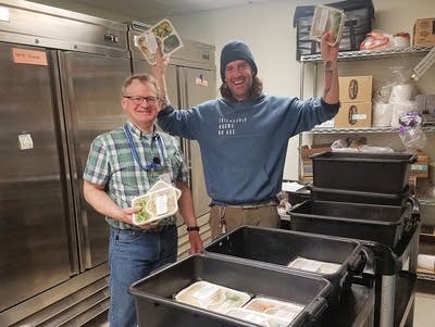 Two people stand in front of bins filled with packaged meals.