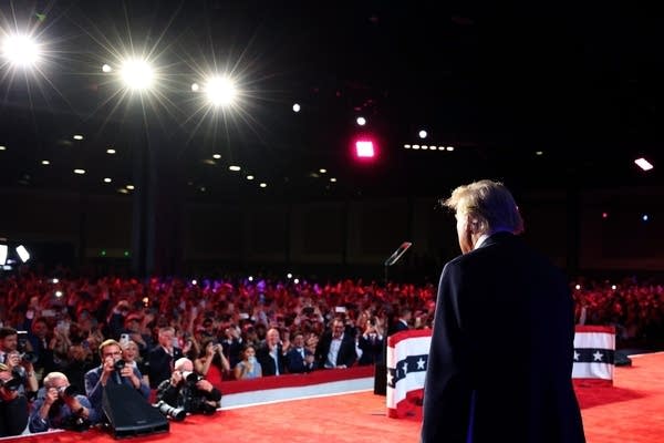 The back of former President Donald Trump as he walks onto a red stage in front of a sea of people.