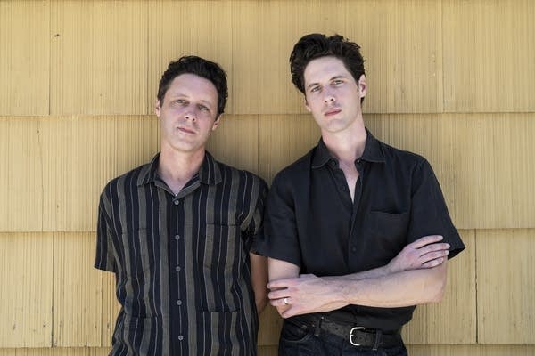 Two men stand in front of an exterior wall clad in cedar shakes