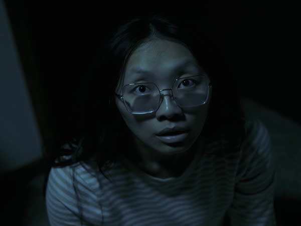 A person with glasses looks up toward the camera in the dark.