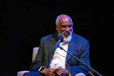 A man and woman sit in chairs on a stage, speaking into microphones.