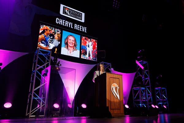 A woman speaks at a podium onstage in front of purple lights and a screen of photos of her.