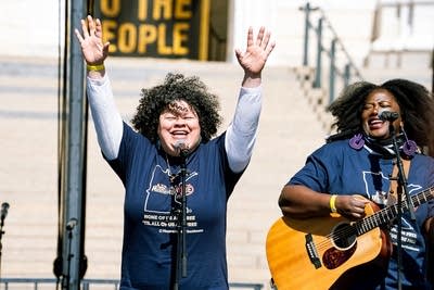 Two musicians perform together on an outdoor stage at a large rally