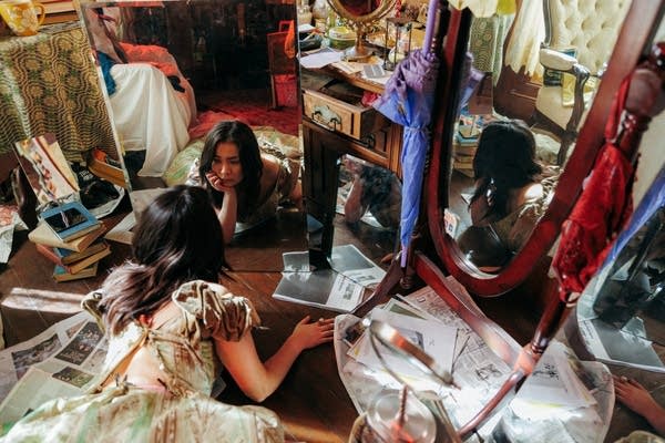 A woman reclines on the floor in front of a mirror in a room cluttered with bric-a-brac
