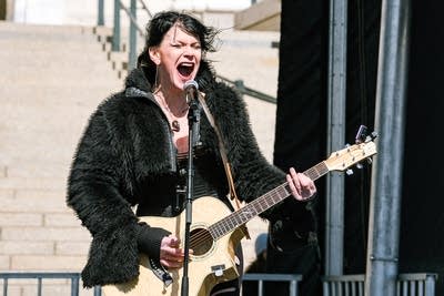 A musician sings and plays guitar on an outdoor stage at a large rally