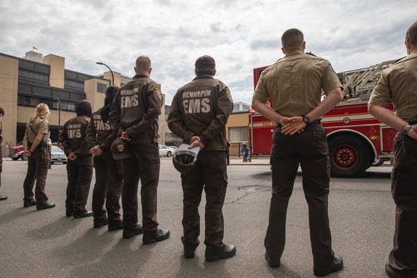 People in brown EMS jackets stand with their hands behind their backs. 