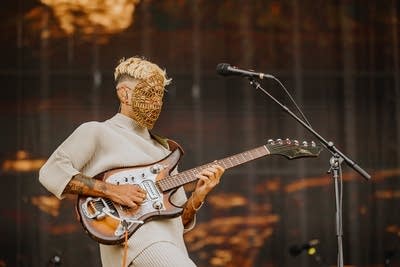 A musician in a golden mask plays guitar on a large outdoor stage