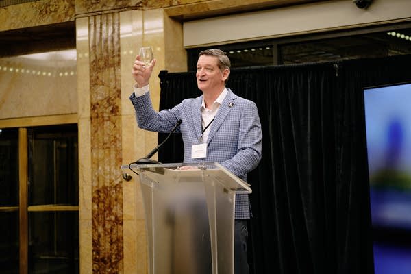 A man speaks at a podium and holds a small glass up.