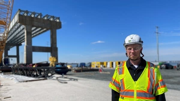 A man in a helmet and a yellow vest stands in front of bridge remains.