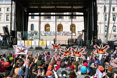 A man with a guitar waves to the audience from an outdoor stage at a large rally