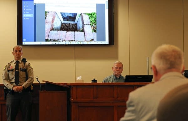 A witness sits in the stand with a screen behind him.