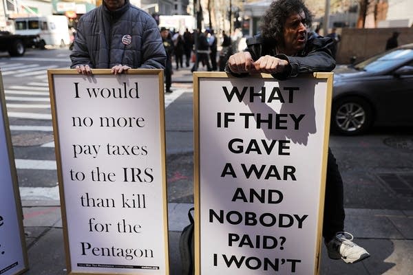 People holds sign saying "I would no more pay taxes to the IRS than kill for the Pentagon" and "What if they gave a war and nobody paid? I won't"