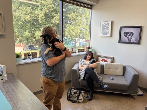 Two women hold puppies. One stands while another sits on a couch.