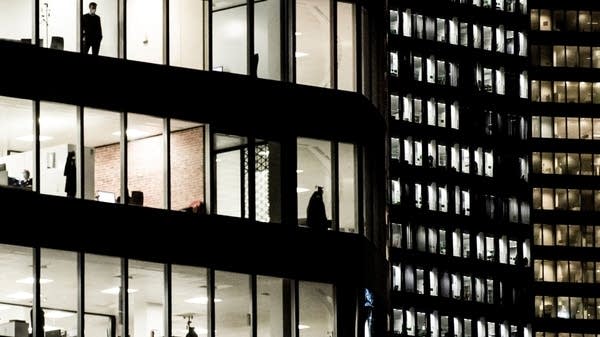 The exterior of two office buildings, lit on the inside, showing desks and people in silhouette.