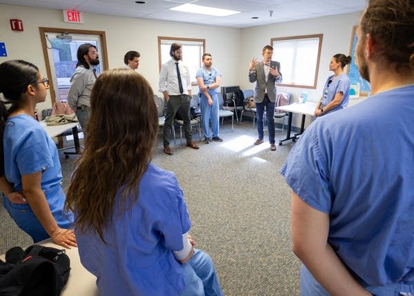 A large group of people wearing scrubs stand in a circle, listening to one man speak.