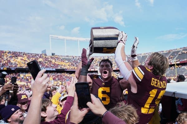 A college football player lifts up a jug-shaped trophy in the middle of a crowd.