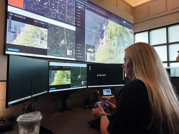 A woman looks at a wide array of screens while holding a controller.