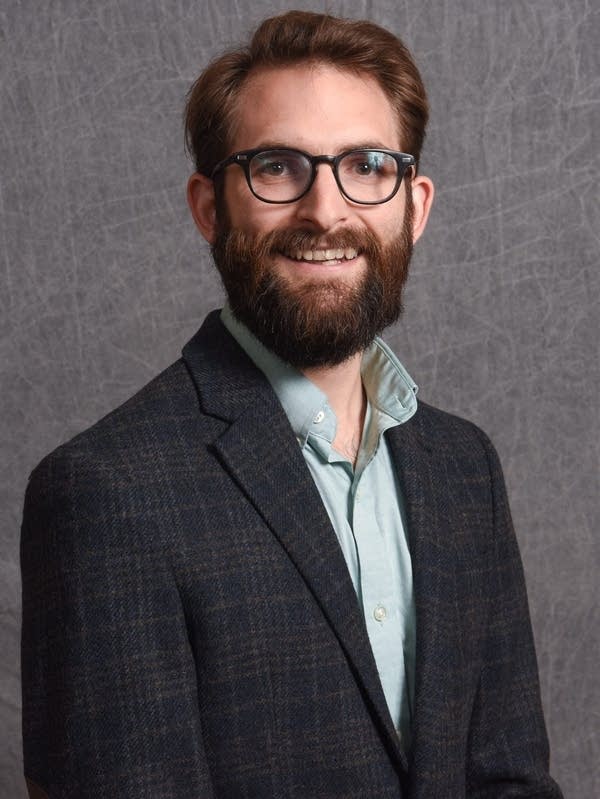 Brad Reaves smiles for his headshot in front of a grey background wearing a plaid blazer.