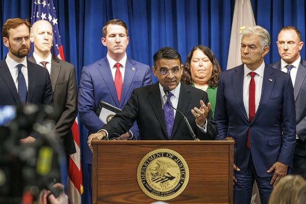 A man in a black suit speaks into a microphone at a podium while flanked by other officials.