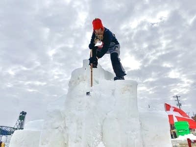 A person carves a snow sculpture while standing on top of it.