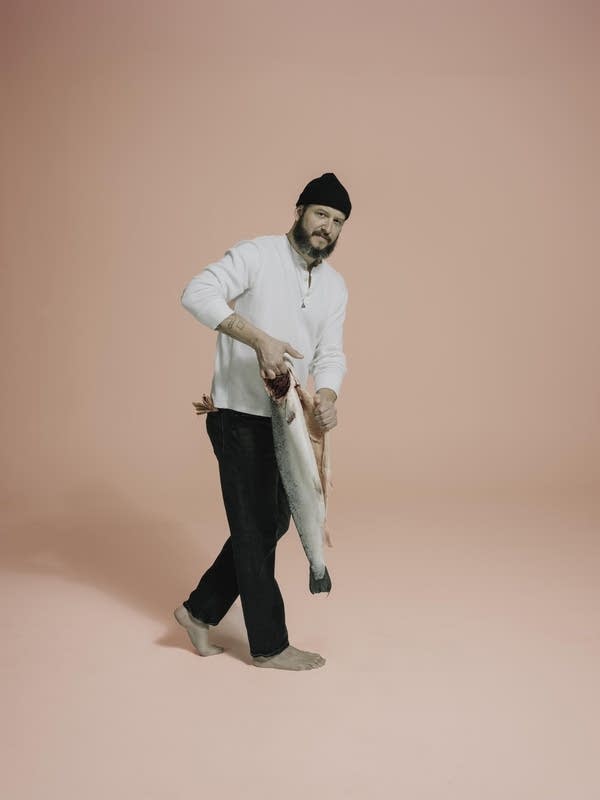 A bearded man in a woolly cap carries a freshly gutted and headed fish
