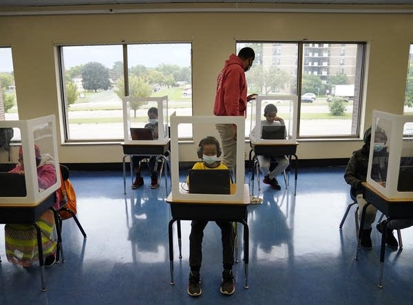 Children sit behind plastic barriers at desks spaced apart in a classroom.