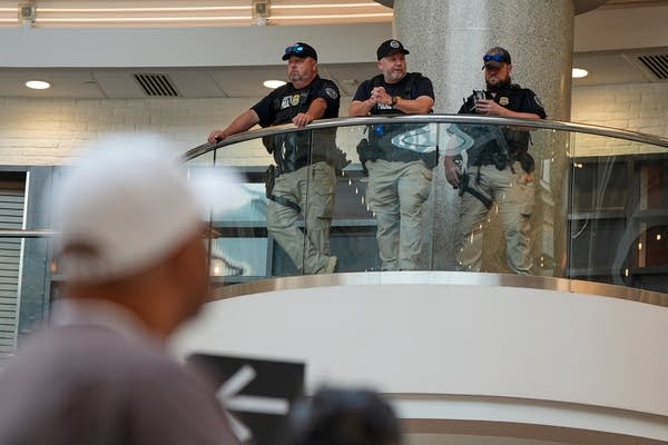 Three federal agents lean against a railing overlooking a crowd of travelers. One is on his phone.