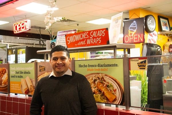 A man poses for a photo in front of the counter for his restaurants. There are bright red lights and menu photos of food.
