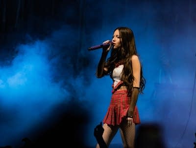 A woman in a plaid skirt and white shirt holds a microphone to her mouth