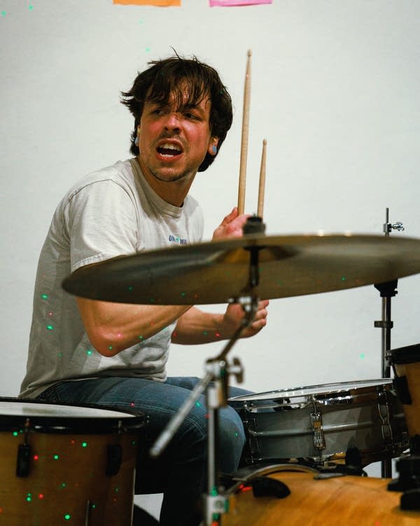 A man wearing a white shirt plays a drum kit.